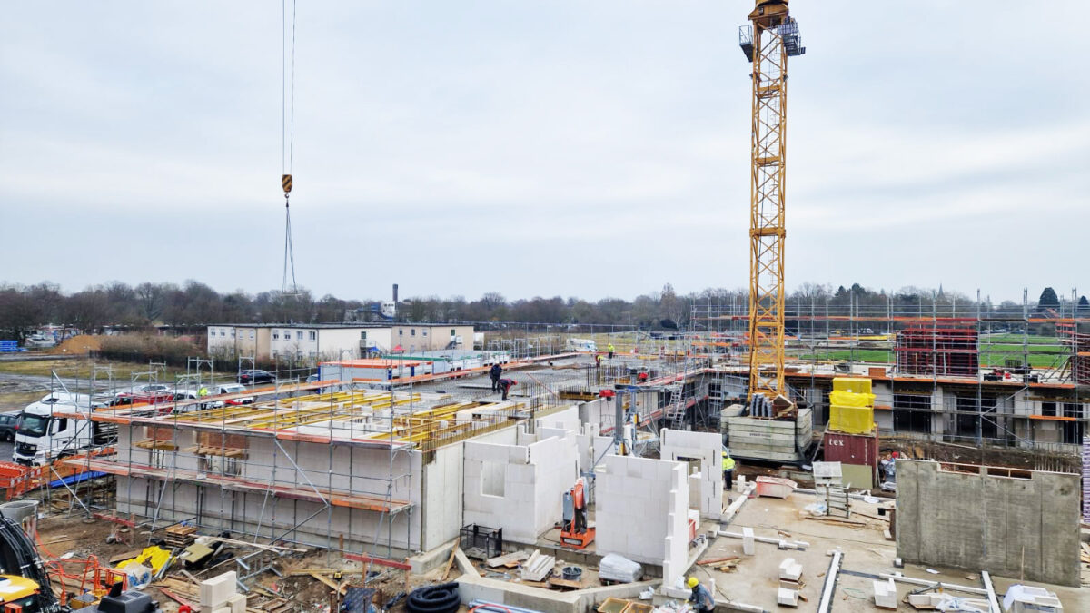 Blick auf Haus Thomas, im Hintergrund Flüchtlingsunterkunft und Schwimmbad: Die Decke eines Traktes des Erdgeschosses ist betoniert, die ersten Wände im 1. Obergeschoss werden hergestellt. Im benachbarten Trakt werden Deckenplatten im Erdgeschoss verlegt. Im Bauteil daneben laufen die Mauerwerksarbeiten.